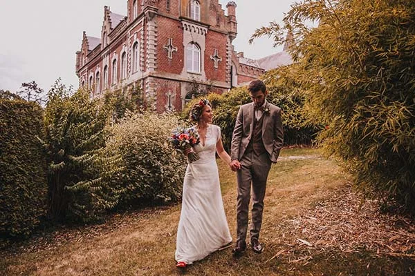 Jeunes mariés marchant main dans la main devant un château belge, ambiance champêtre et romantique.