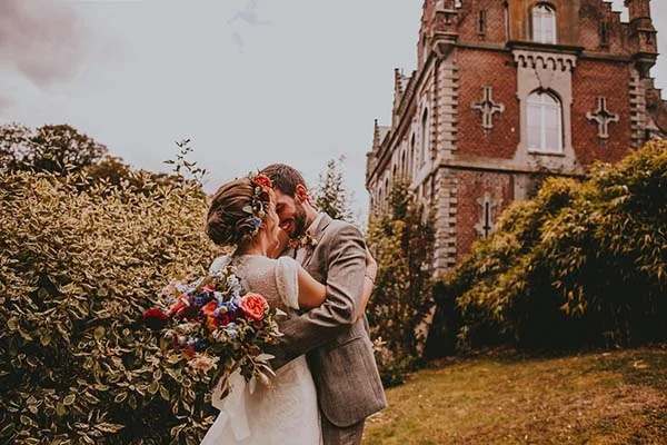 Jeunes mariés s'enlaçant devant un château en briques, entourés de végétation et d'un bouquet coloré.