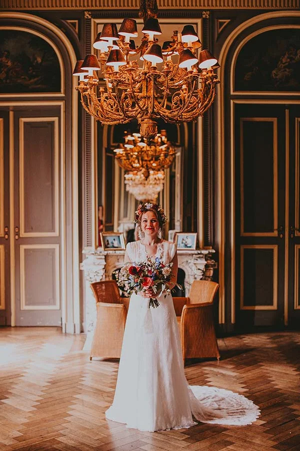 Mariée en robe blanche avec couronne et bouquet colorés, posant dans un salon classique sous un lustre doré.