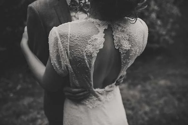 Gros plan en noir et blanc du dos d'une mariée en robe de dentelle, enlacée par son époux dans un cadre naturel.
