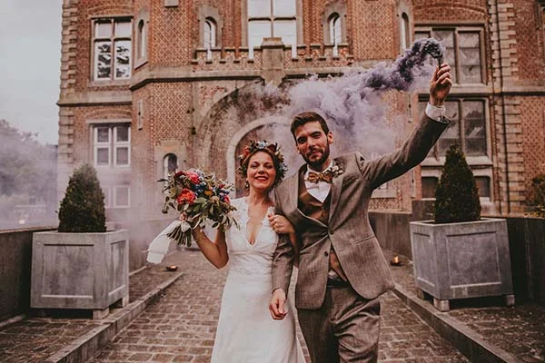 Jeunes mariés souriants devant un château belge, avec fumigène violet et bouquet coloré dans une ambiance festive.