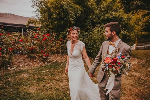 Jeunes mariés souriants main dans la main dans un jardin champêtre, avec bouquet coloré et couronne de fleurs.
