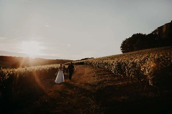 Jeunes mariés marchant main dans la main dans un vignoble occitan au coucher du soleil, ambiance romantique et dorée.