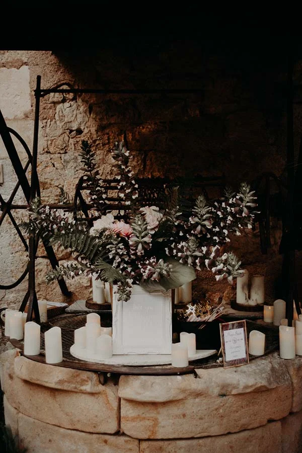 Composition florale blanche et feuillage dans un vase blanc entouré de bougies sur un puits en pierre rustique.