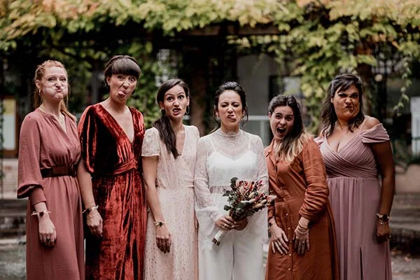 Six femmes en robes élégantes aux tons terracotta et blanc font des grimaces amusantes lors d'un mariage en extérieur.