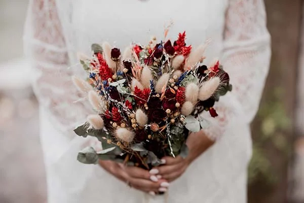 Mariée tenant un bouquet champêtre aux tons rouge profond, crème et eucalyptus pour un mariage romantique.