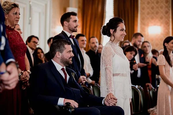 Une mariée en robe blanche et son époux en costume bleu sourient entourés de leurs invités lors d'une cérémonie.