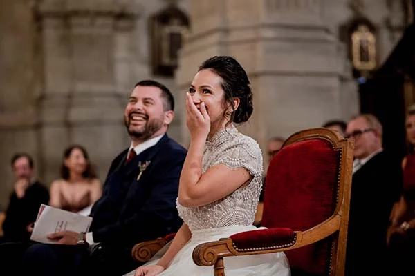 Jeunes mariés riant avec complicité, elle en robe blanche perlée et lui en costume bleu, dans un cadre solennel.