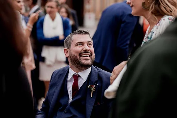 Un marié souriant en costume bleu marine et cravate rouge lors d'une réception de mariage chaleureuse.