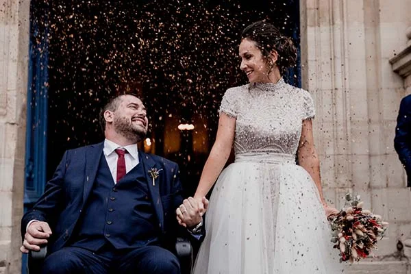 Jeunes mariés souriants sous une pluie de confettis dorés, elle en robe blanche et lui en costume bleu marine.