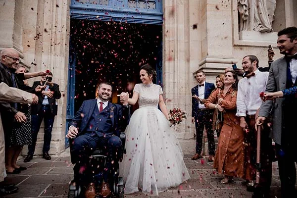 Jeunes mariés sortant d'une église sous une pluie de pétales rouges, entourés d'invités dans une ambiance festive.
