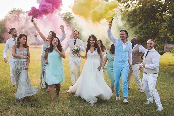 Jeunes mariés et invités en tenues claires célébrant joyeusement avec des fumigènes colorés dans un champ.