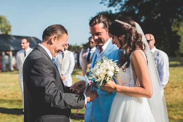 Un marié en costume bleu et une mariée en robe blanche échangent leurs vœux lors d'une cérémonie champêtre et lumineuse.