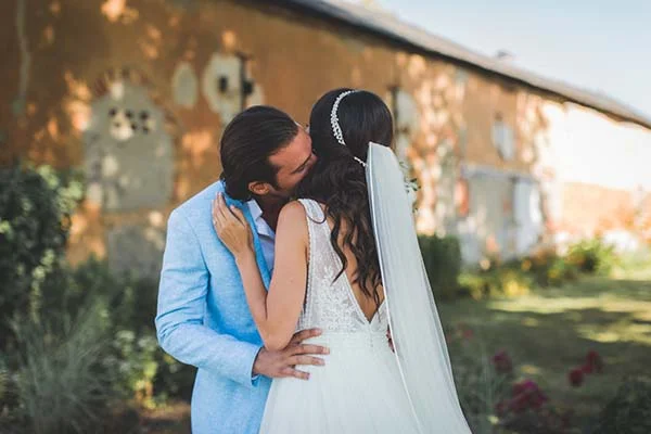 Jeune marié en costume bleu et mariée en robe blanche s'embrassant devant un mur rustique, ambiance chic et champêtre.