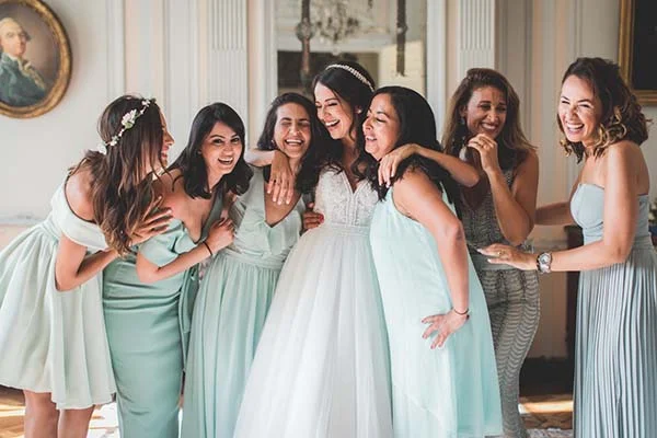 Une mariée en robe blanche rit entourée de ses demoiselles d'honneur en robes vert menthe dans un intérieur élégant.