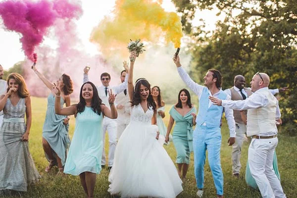 Mariés et invités célébrant joyeusement en plein air avec des fumigènes roses et jaunes dans un style champêtre.