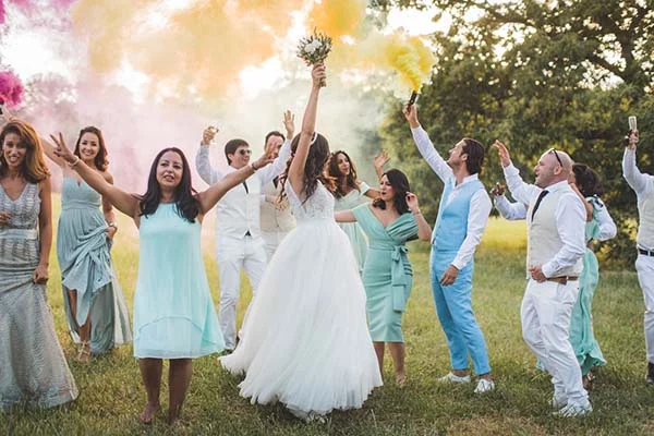 Une mariée et ses invités célèbrent joyeusement en plein air sous des fumigènes colorés dans un style chic et champêtre.