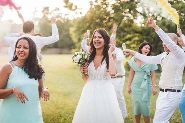 Une mariée rayonnante en robe blanche rit entourée de ses invités lors d'une fête champêtre et festive en extérieur.