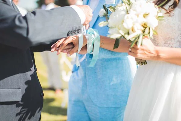 Gros plan sur les mains d'un couple marié, avec un bouquet blanc et des tenues élégantes bleu ciel et blanc.