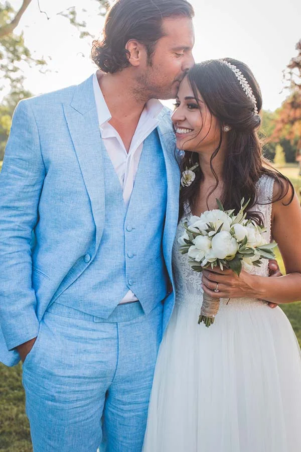 Un couple de mariés s'embrasse tendrement, elle en robe blanche et lui en costume bleu clair, dans un cadre champêtre.