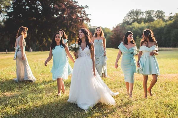 Une mariée rayonnante et ses demoiselles d'honneur en robes bleu pastel marchent dans un champ ensoleillé.