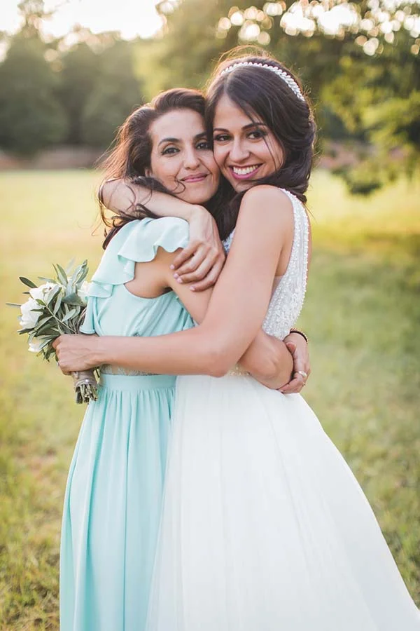 Deux femmes souriantes s'enlaçant tendrement, l'une en robe blanche et l'autre en robe bleu ciel, dans un cadre champêtre.