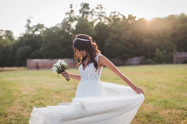 Une mariée en robe blanche tournoyant avec son bouquet dans un champ ensoleillé à l'ambiance champêtre.