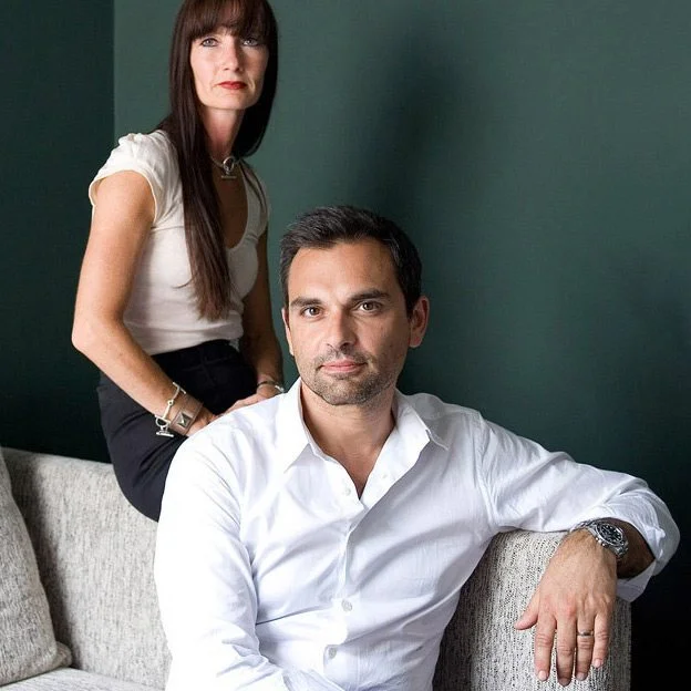Un couple élégant dans des costumes blancs, assis sur un canapé avec un fond vert profond, émettant un sentiment de