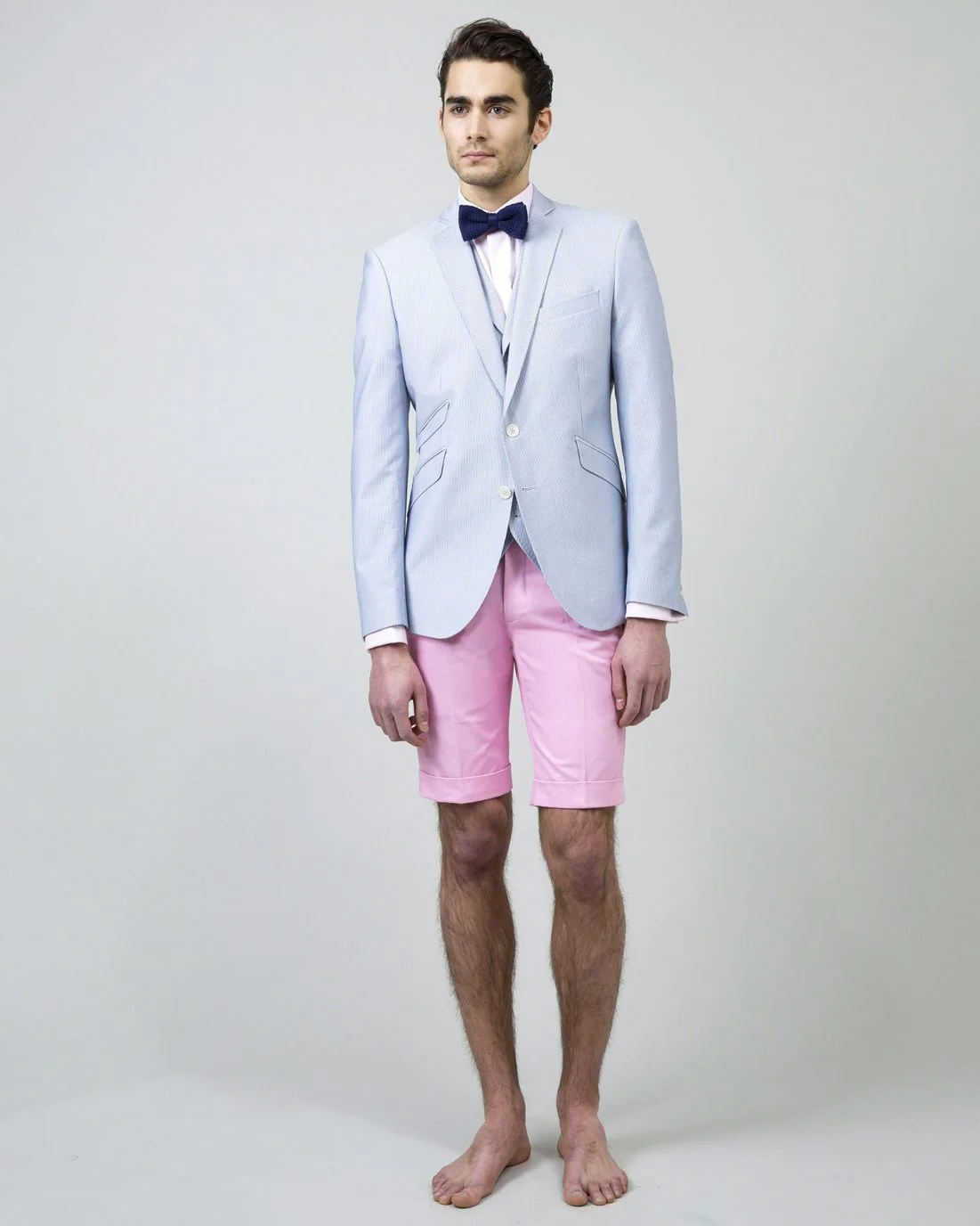 Un jeune homme habillé d'un costume bleu pâle et de shorts rose, prêt pour une cérémonie de mariage décontracté et raffiné.