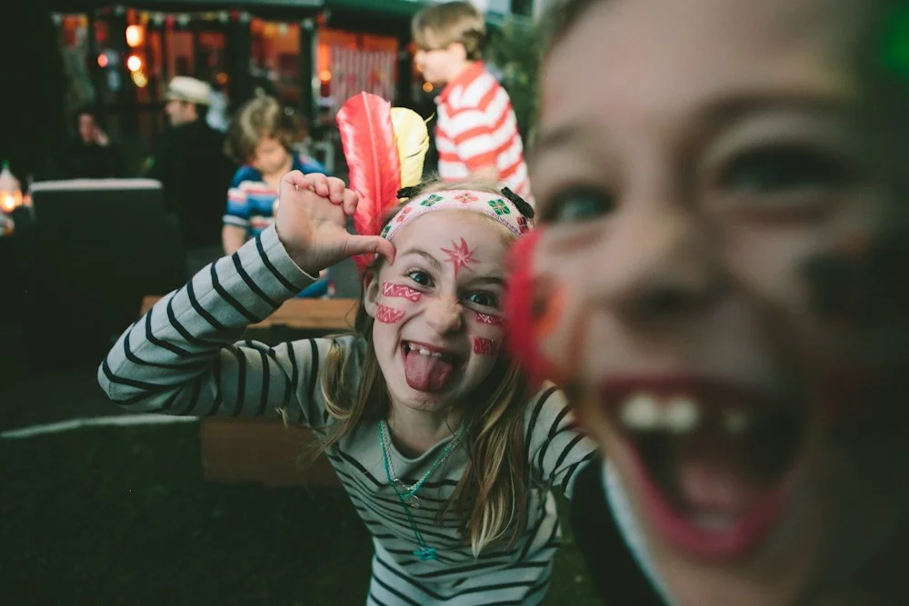Une jeune fille joue avec des poudres de riz colorées, son visage peint, dans un décor de fête animé.