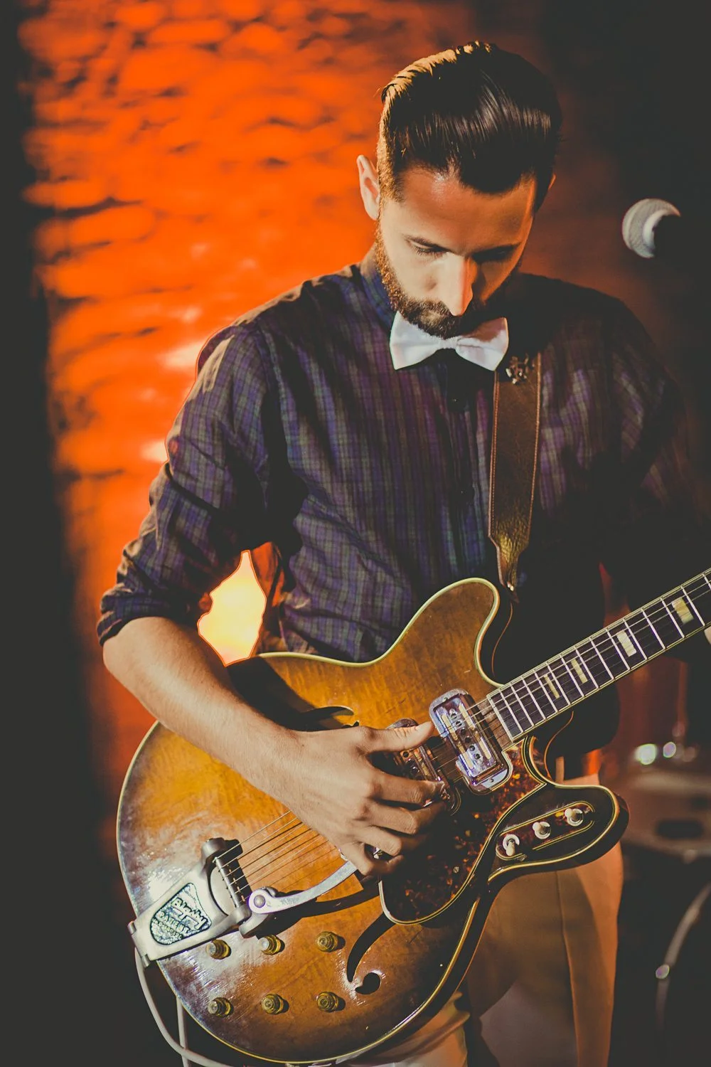 Un homme à la coiffure soignée et un nœud blanc joue de guitare avec passion dans un décor chaud et accueillant.