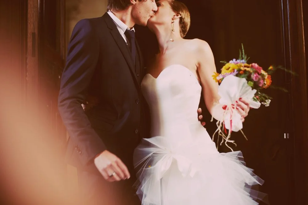 Un couple de mariés partageant un baiser tendre, l'homme en costume sombre et la femme en robe blanche avec un bouquet coloré.