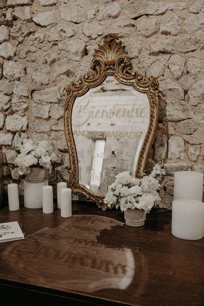 Miroir doré avec message de bienvenue, entouré de fleurs blanches et bougies sur un meuble rustique devant un mur de pierre.