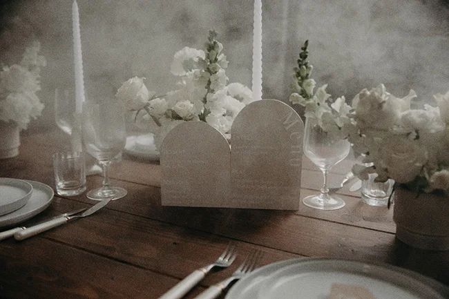 Table de mariage chic avec fleurs blanches, bougies et vaisselle épurée sur un bois sombre dans une ambiance vaporeuse.