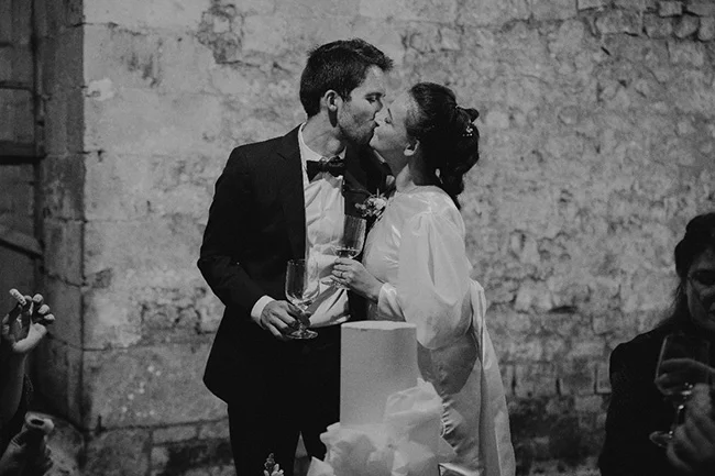 Jeunes mariés s'embrassant avec des coupes de champagne devant un gâteau, photo en noir et blanc au style chic.