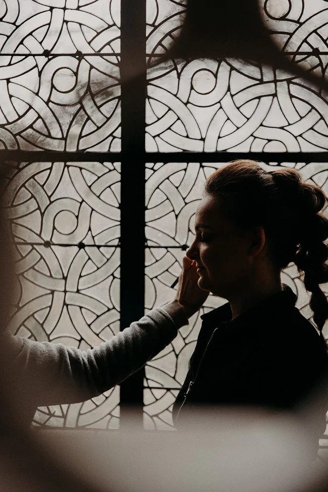 Silhouette d'une mariée se faisant maquiller devant un vitrail aux motifs entrelacés, ambiance douce et contrastée.