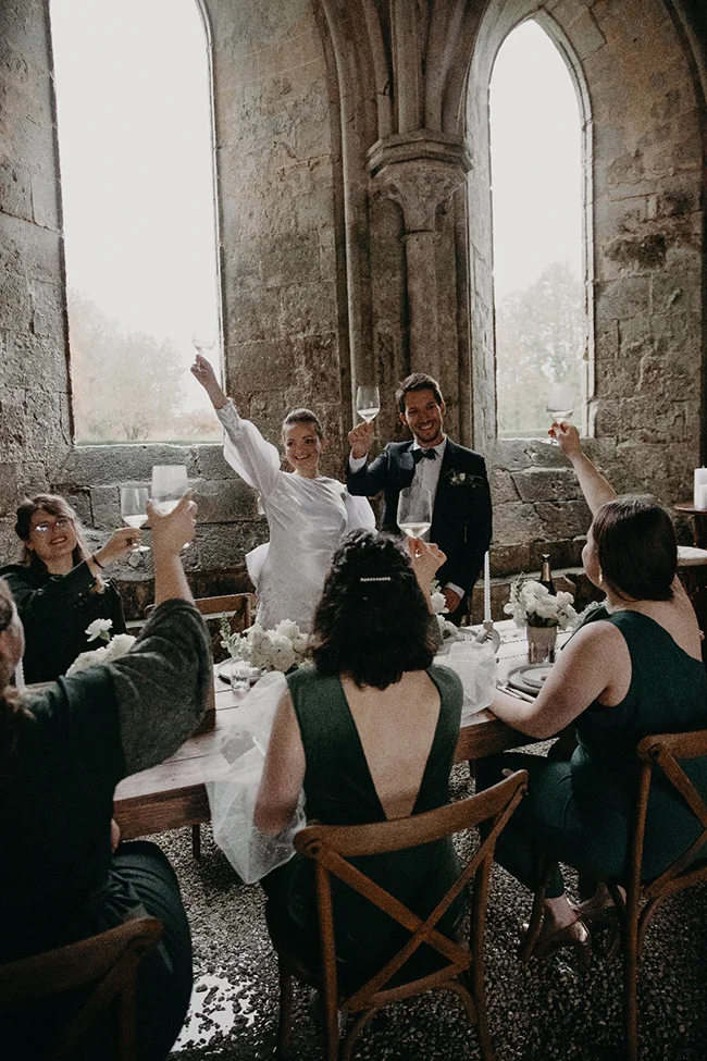 Jeunes mariés et invités portant un toast avec élégance dans un cadre architectural en pierre aux tons neutres.