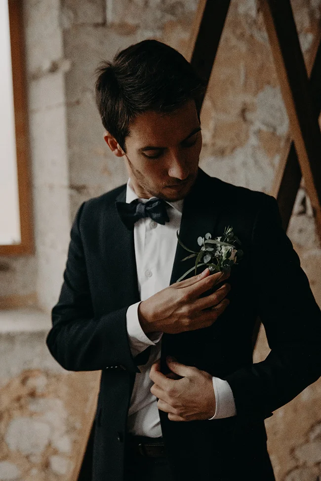 Homme en smoking noir et nœud papillon ajustant sa boutonnière végétale dans un cadre rustique et élégant.