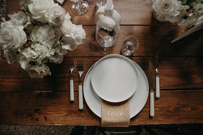 Dressage de table chic sur bois brut avec assiettes blanches, fleurs blanches et marque-place au nom de Félicie.