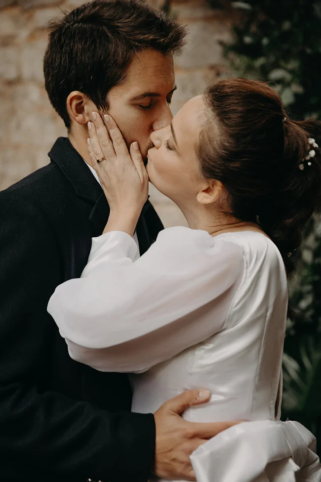 Jeunes mariés s'embrassant tendrement, elle en robe blanche et lui en costume noir, dans un cadre naturel et chic.