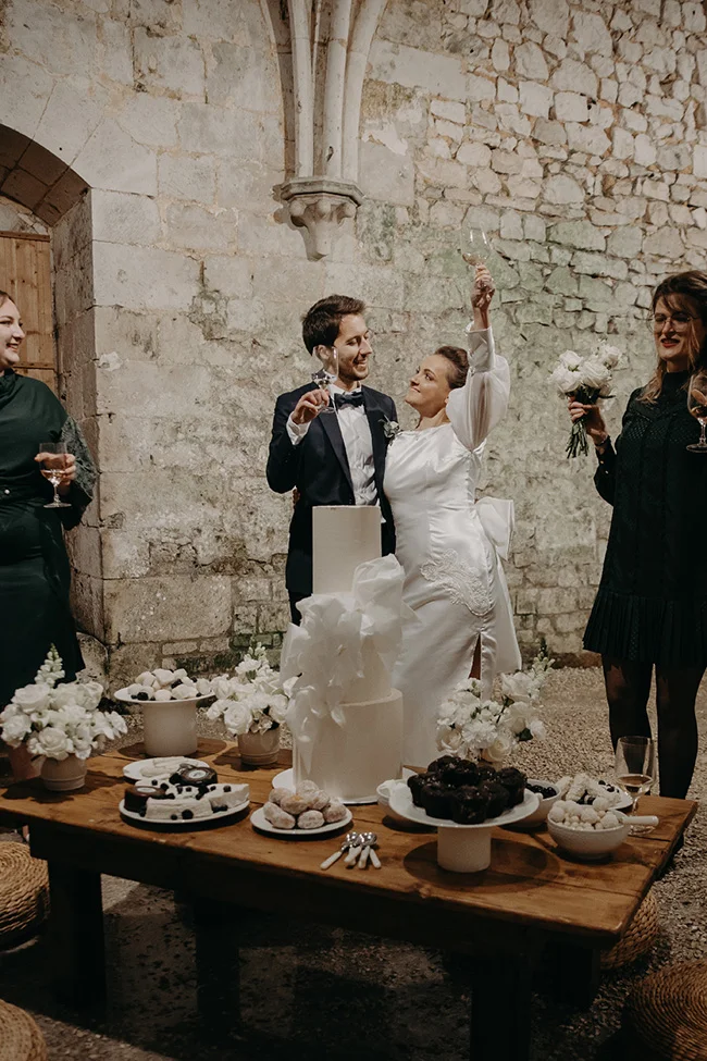 Jeunes mariés célébrant avec champagne devant un gâteau blanc et des fleurs, dans un cadre rustique et chic.