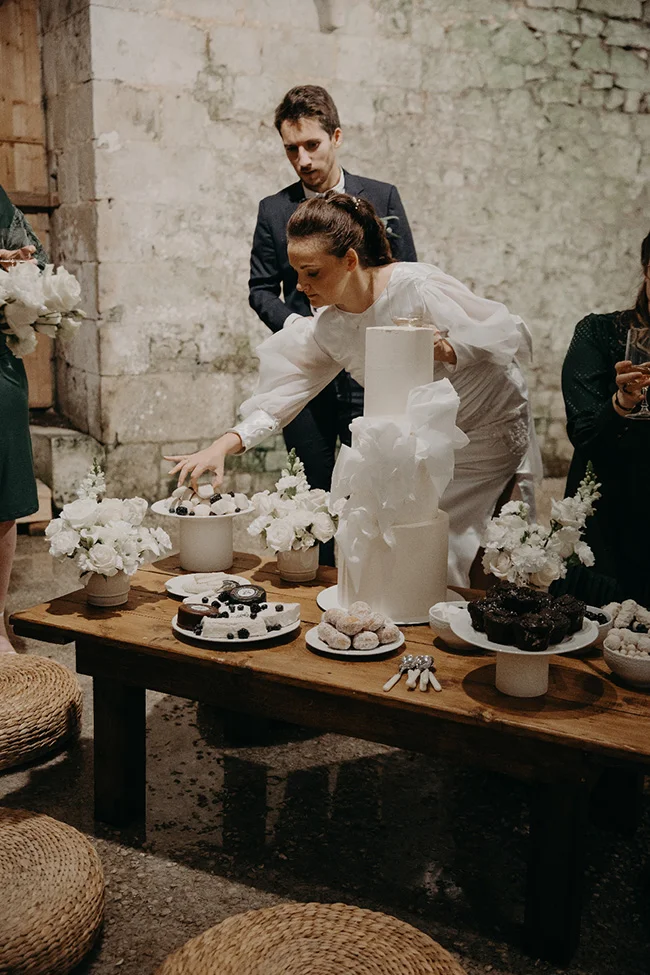 Une mariée en robe blanche sert des mignardises sur une table en bois lors d'une réception chic et épurée.