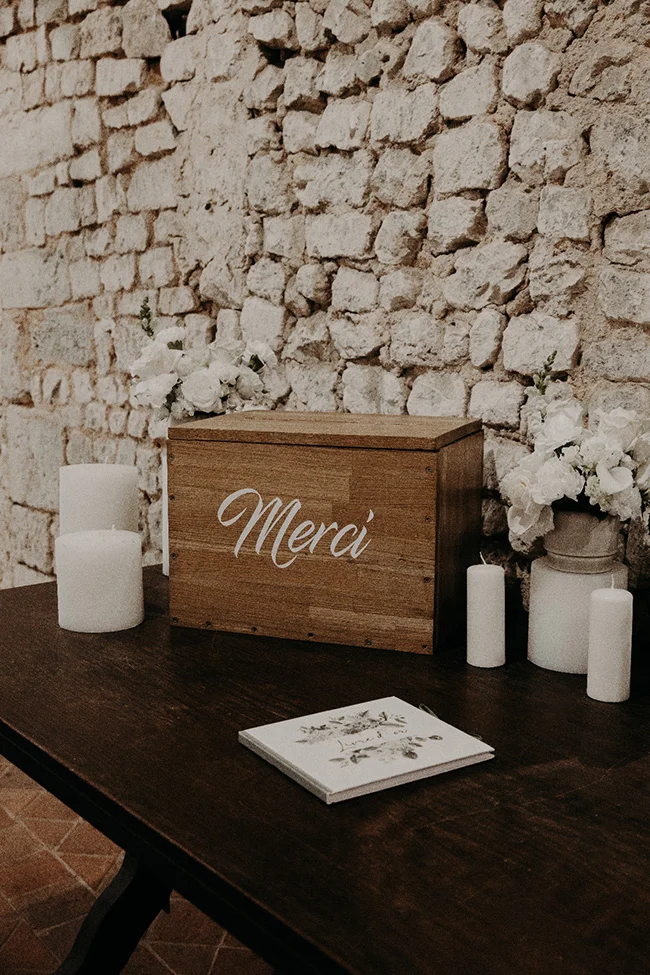 Urne en bois marquée Merci sur une table sombre avec bougies blanches et fleurs, devant un mur en pierres claires.
