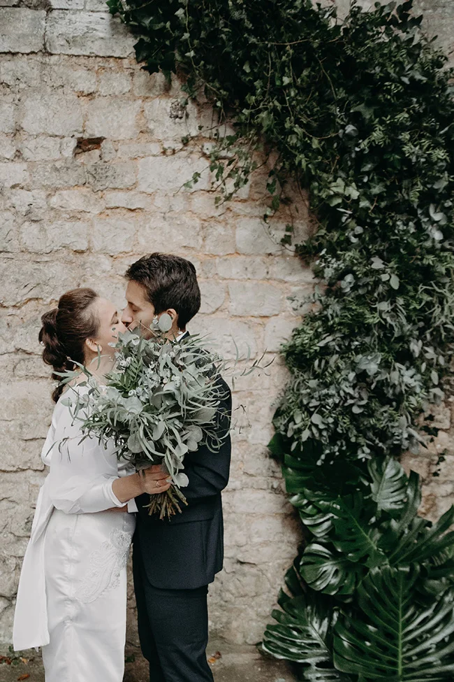 Jeune couple marié s'embrassant tendrement devant un mur de pierre recouvert de lierre avec un grand bouquet végétal.