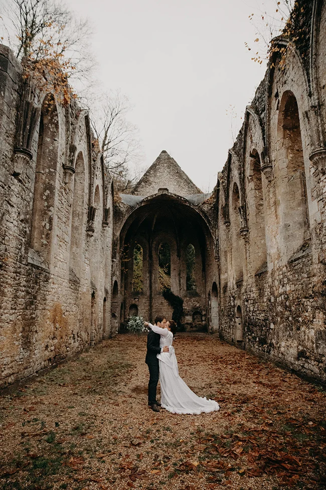 Couple de mariés s'embrassant dans les ruines d'une abbaye entourées de feuilles d'automne aux tons chauds.