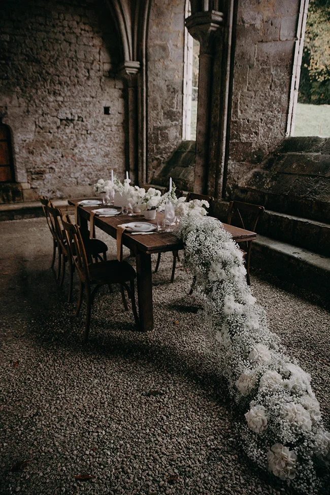 Table de mariage en bois décorée de fleurs blanches et gypsophile dans un cadre rustique en pierre.