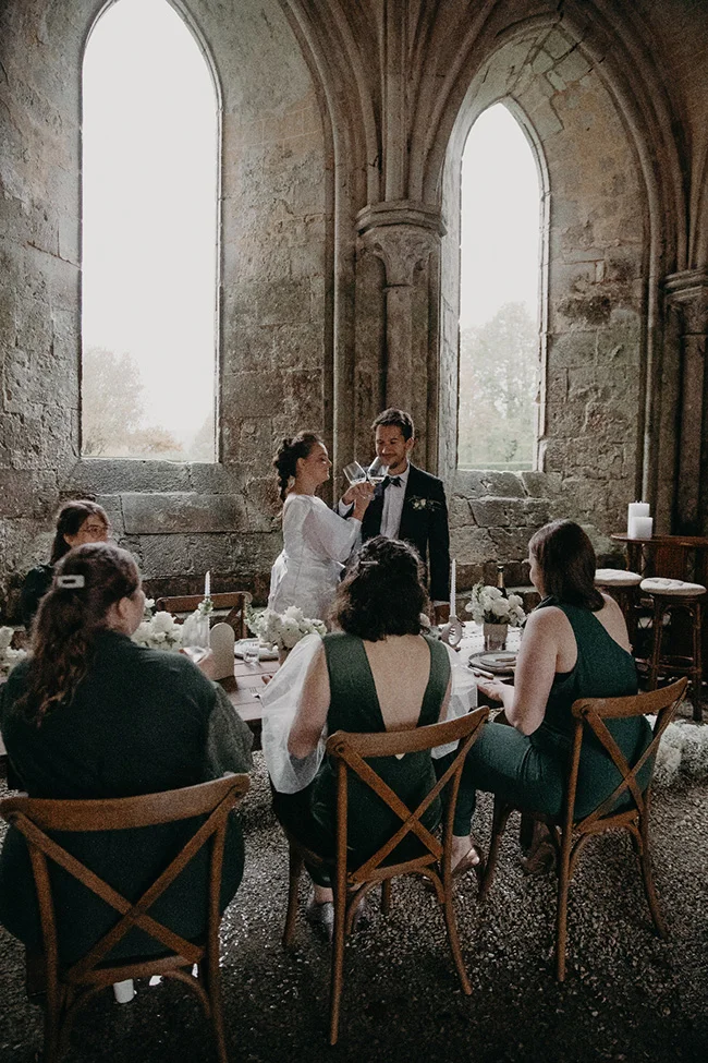 Couple de mariés trinquant lors d'un dîner chic dans une ruine gothique avec des invités en robes vert sapin.