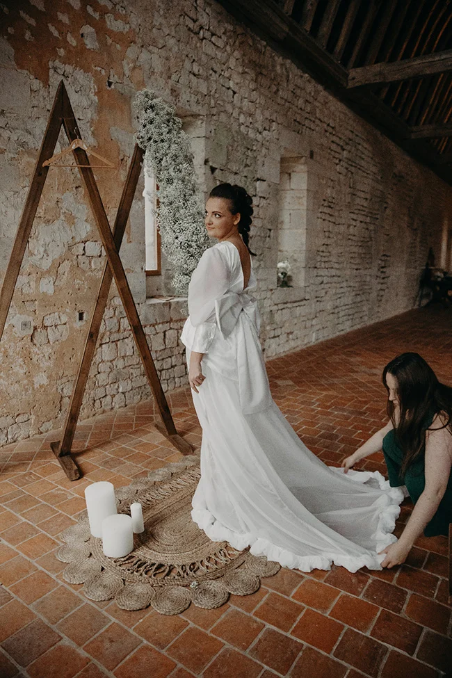 Une mariée en robe blanche élégante pose dans un intérieur rustique en pierre avec des touches végétales.