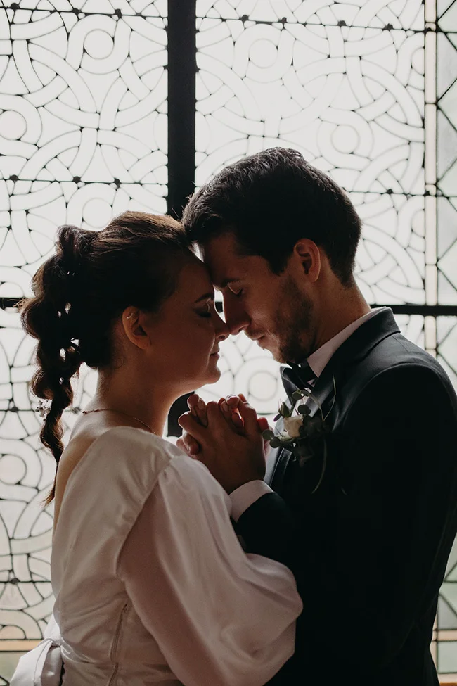 Couple de mariés en silhouette, front contre front, devant une verrière art déco dans une ambiance romantique et chic.