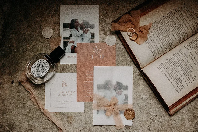Papeterie de mariage beige et blanche, alliances et parfum posés sur un fond minéral aux tons neutres et romantiques.